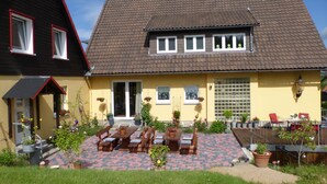 A two-story house with a tiled roof, a front porch with a bench and table, and a small garden area with potted plants and flowers.