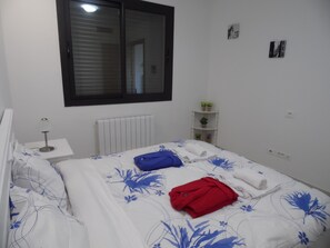 A bedroom with a bed, a bedside table, a radiator, a window, and a shelf with plants.