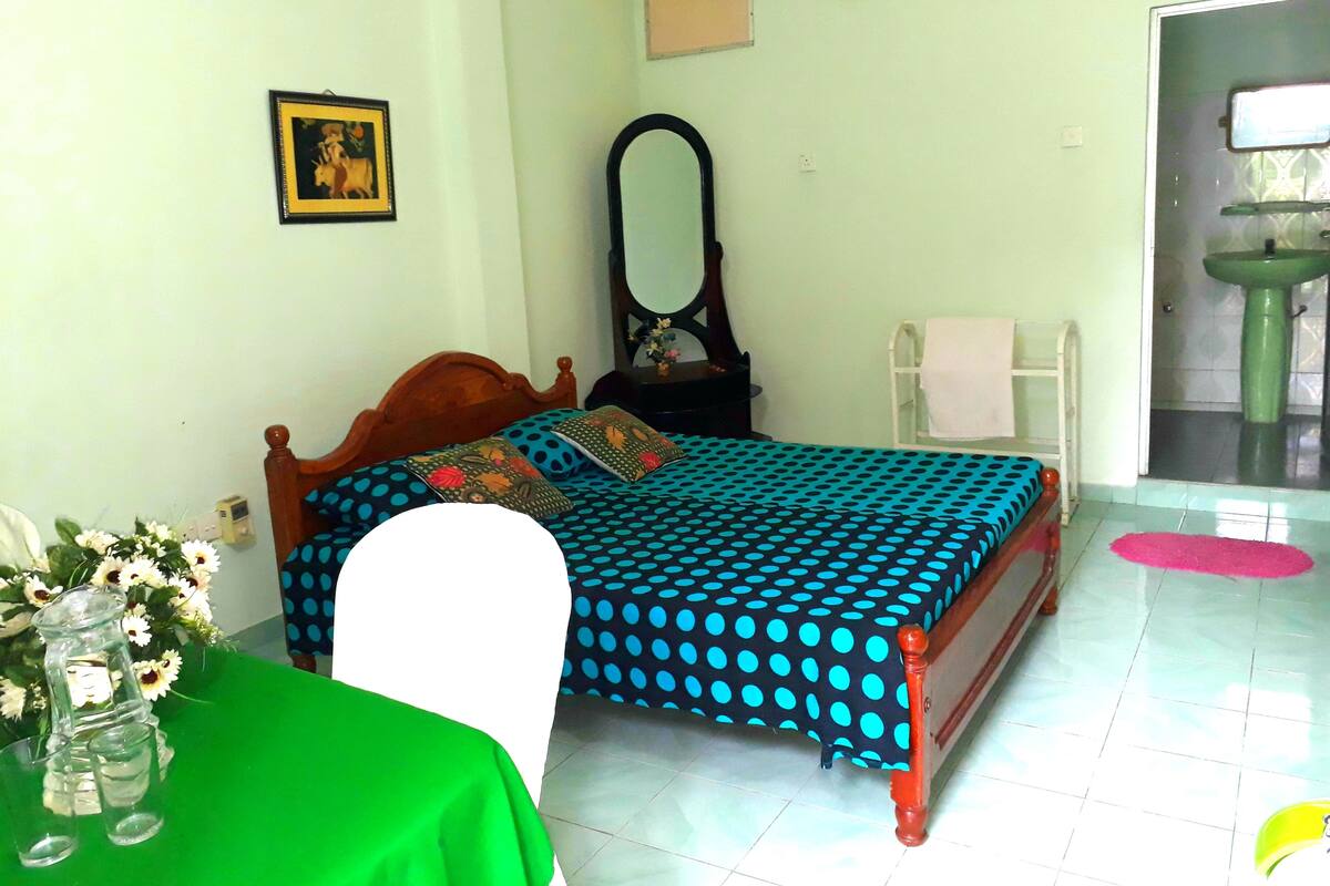 A bedroom with a bed, a mirror, a sink, and a green table with a vase of flowers.