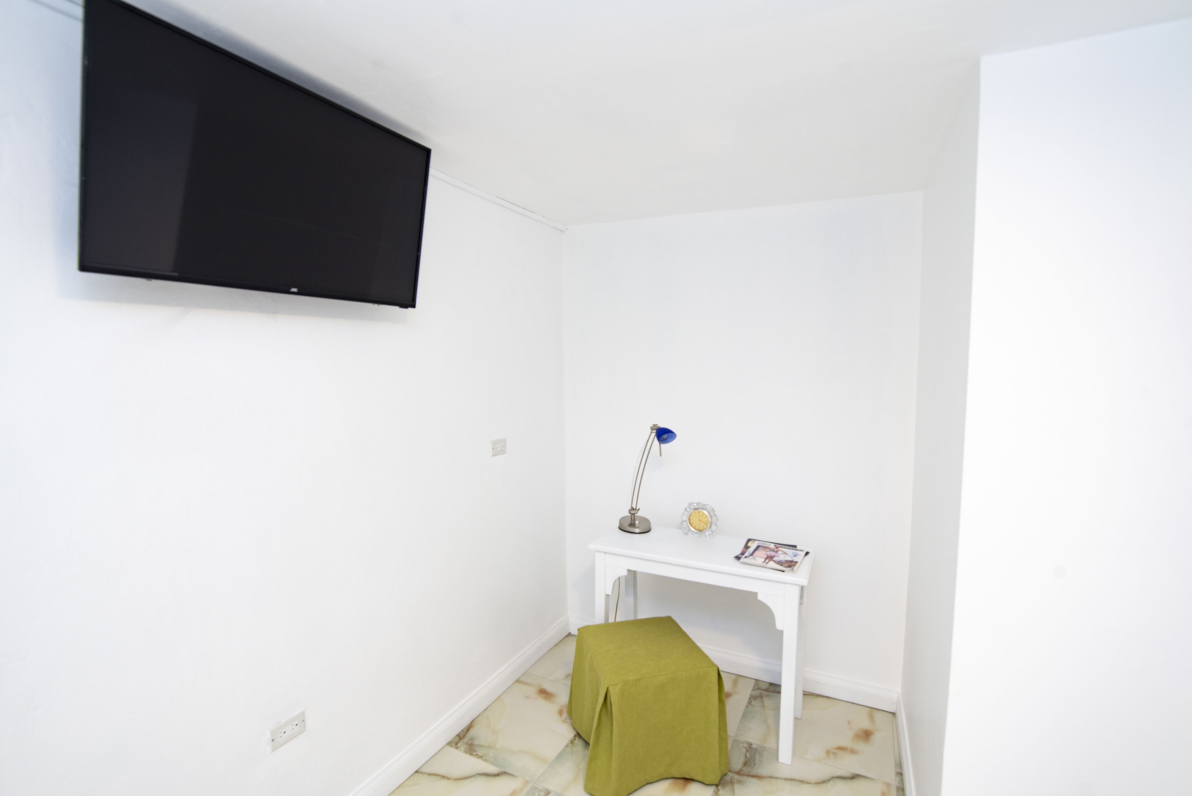 A small, clean room with a white wall, a mounted TV, a white desk with a lamp, and a green cloth on the floor.
