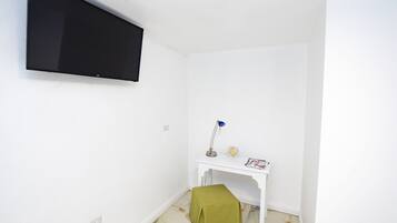 Una stanza piccola e pulita con una parete bianca, una TV a parete, una scrivania bianca con una lampada e un tappeto verde sul pavimento.