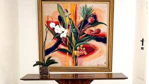 Una habitación con un cuadro enmarcado de flores en la pared, una pequeña planta en maceta sobre una consola y un taburete tapizado en verde.