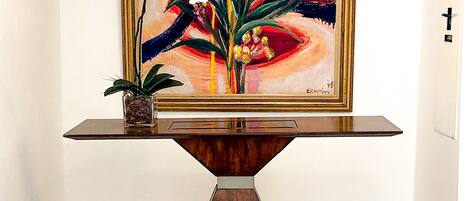 A room with a framed painting of flowers on a wall, a small potted plant on a console table, and a green cloth-covered stool.