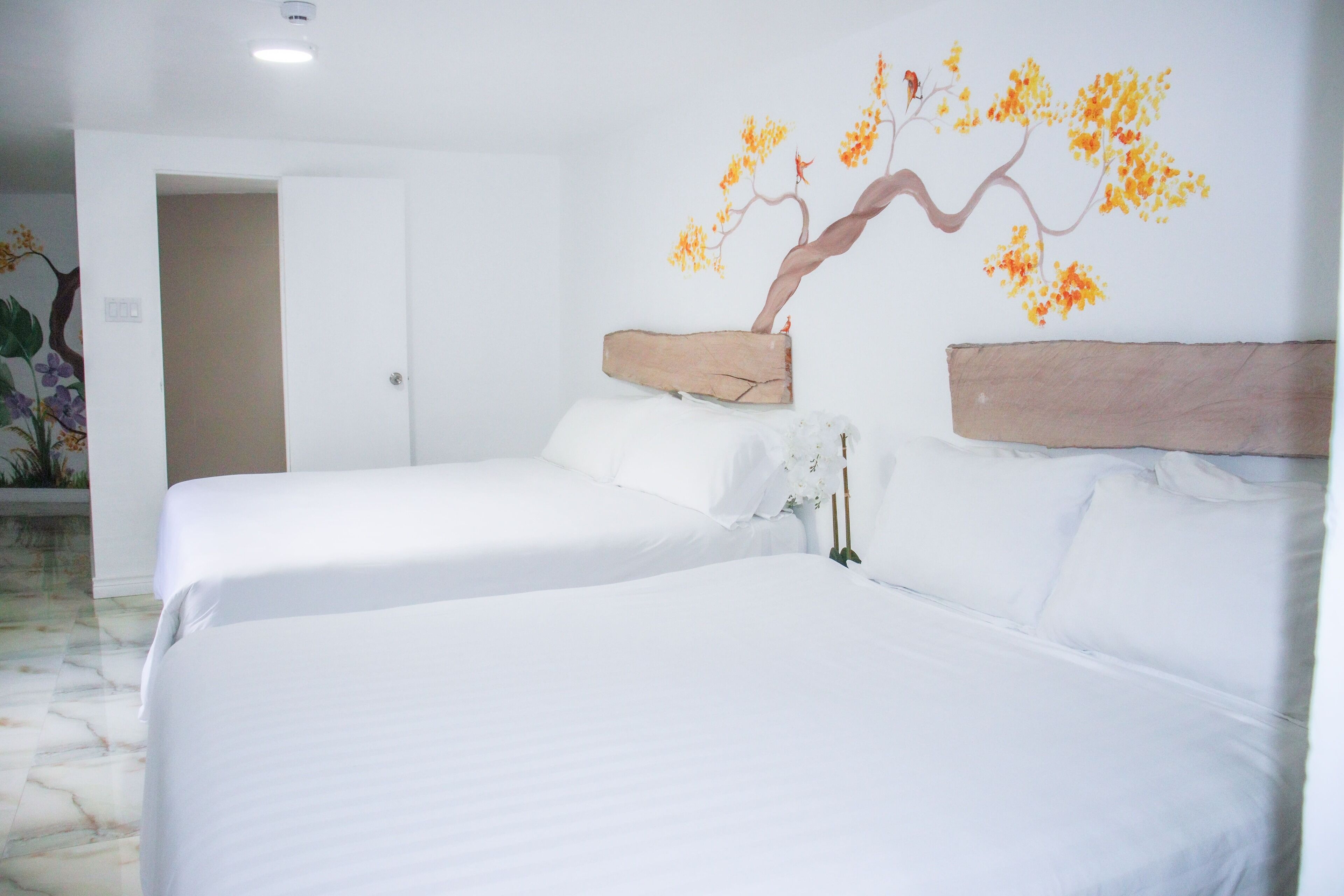 Habitación de hotel con dos camas, ropa de cama blanca, cabecero de madera y un mural de un árbol en la pared.