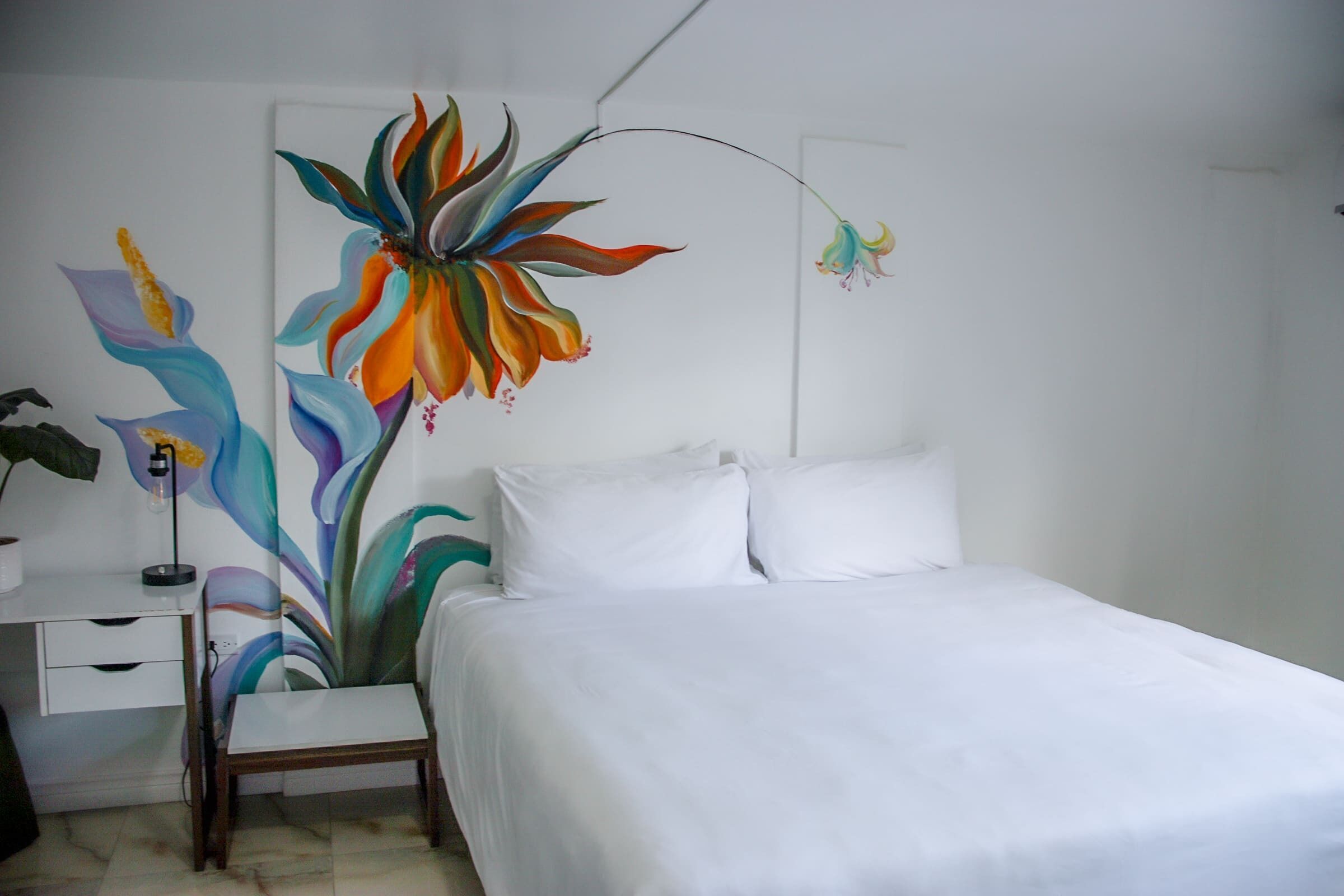A bedroom with a bed, white bedding, a nightstand, a lamp, and a wall painting of a large flower.