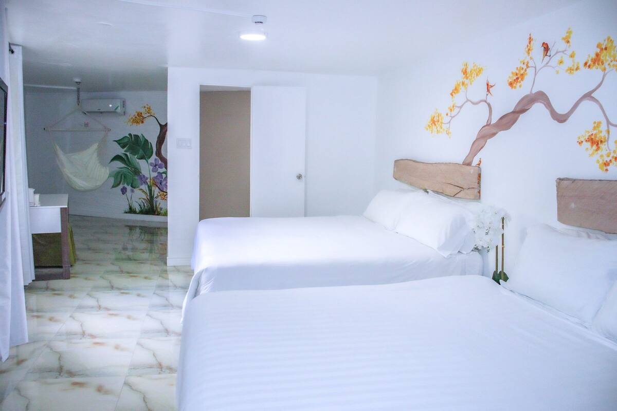 A hotel room with two beds, a mural of a tree with flowers, a hanging chair, and a bathroom with a shower.