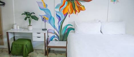 A hotel room with a bed, a desk, a chair, a green stool, and a large floral painting on the wall.