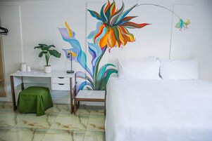 Habitación de hotel con cama, escritorio, silla, taburete verde y un cuadro grande con motivos florales en la pared.