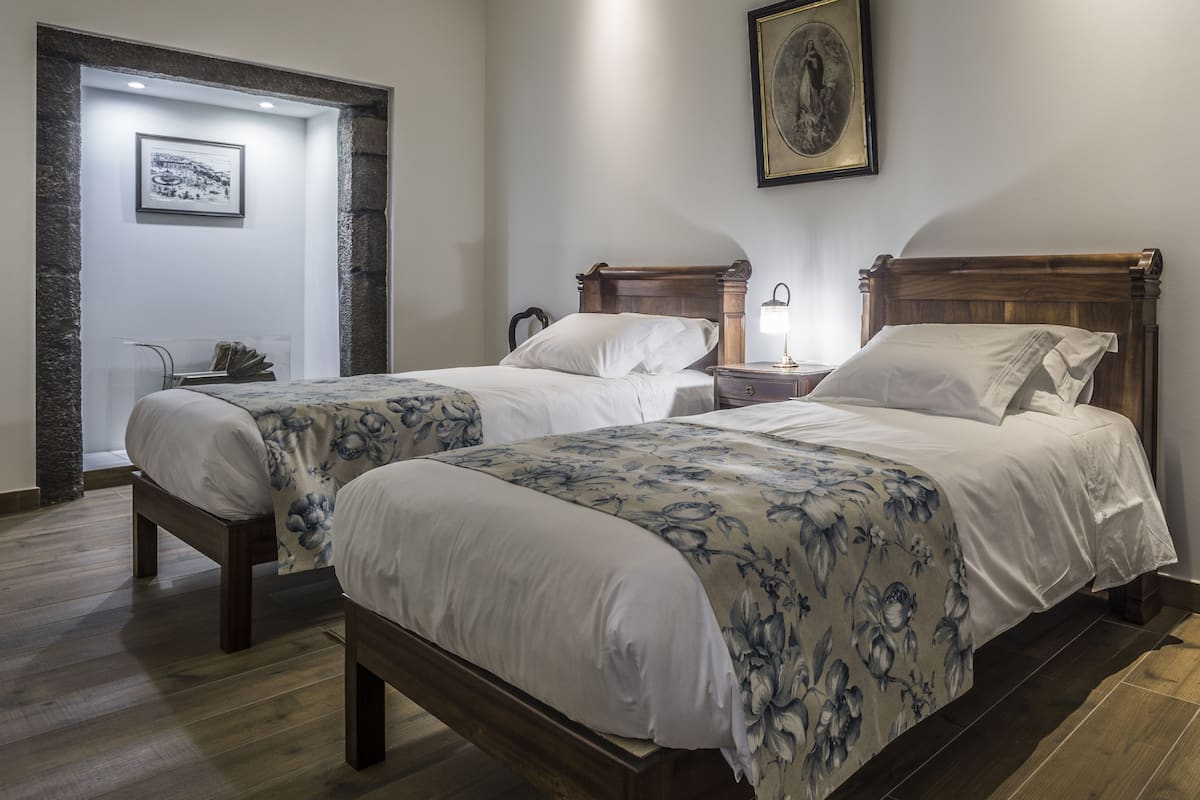 Een hotelkamer met twee houten bedden, een nachtkastje met een lamp, een ingelijst schilderij aan de muur en een stenen zuil.