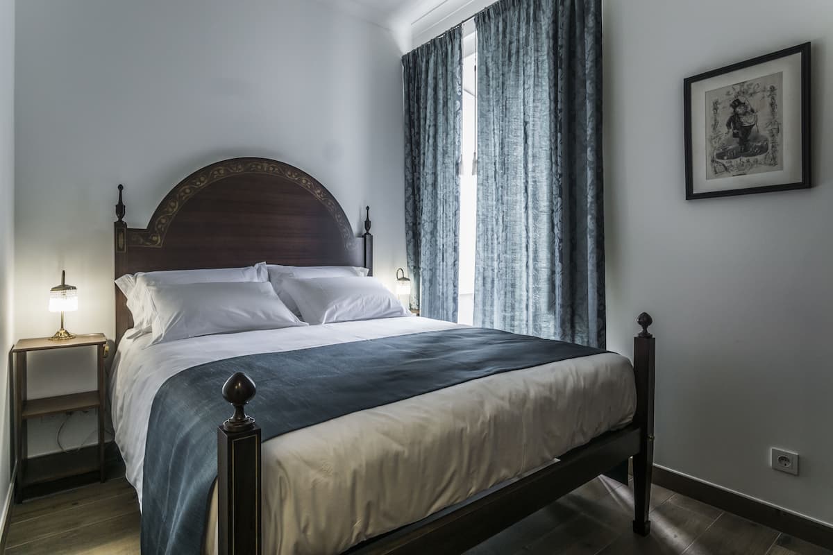 Een net opgemaakt bed met houten hoofdbord, een nachtkastje met lamp en een raam met gordijnen.