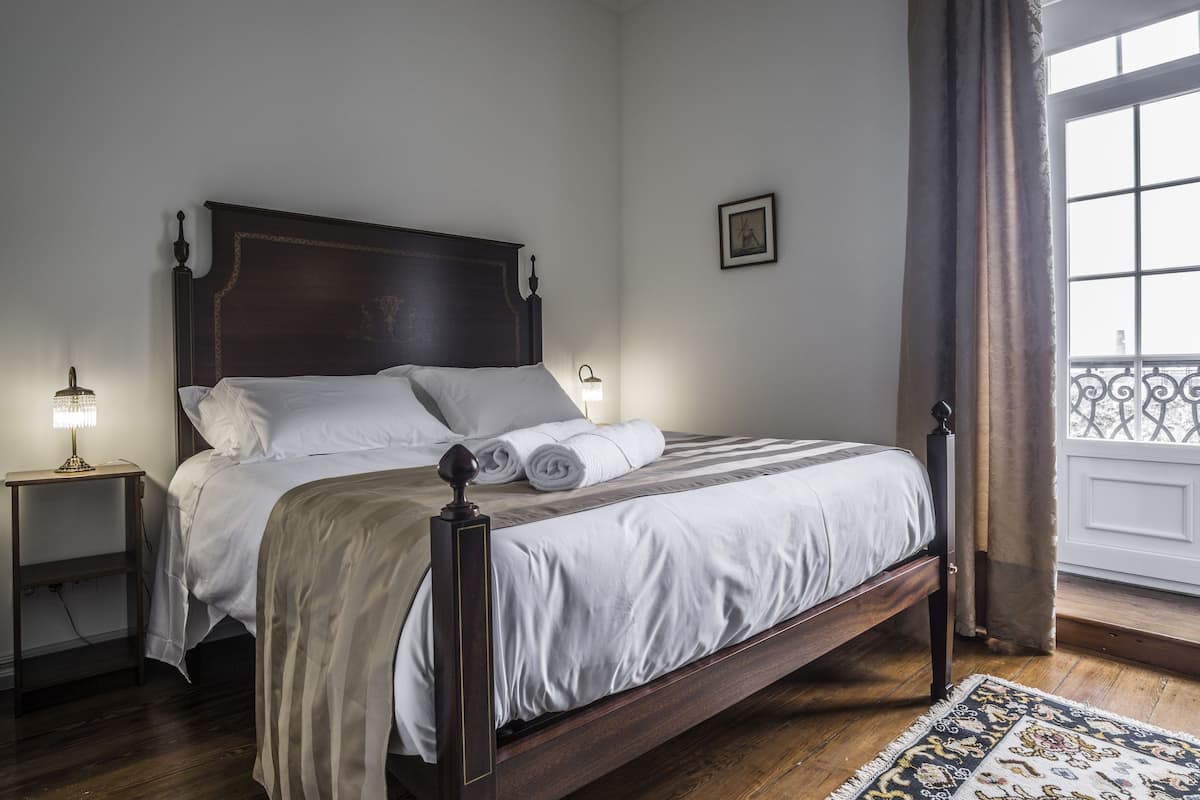 Een slaapkamer met een houten bed, witte lakens, een nachtkastje met een lamp, en uitzicht op het balkon.