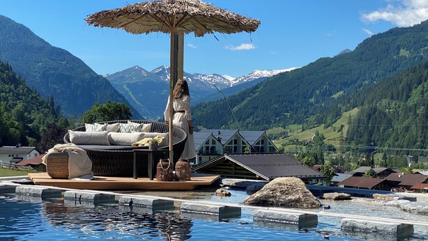 A poolside area with a lounge chair and umbrella, a wooden deck, and a view of mountains and houses. - Aktivhotel Gasteiner Einkehr (Dorfgastein)