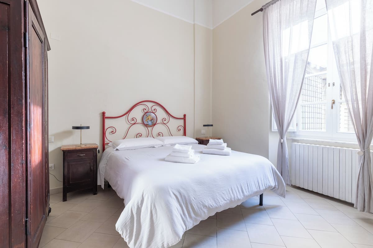 A neatly made bed with white linens, a wooden nightstand with a lamp, and a window with sheer curtains.