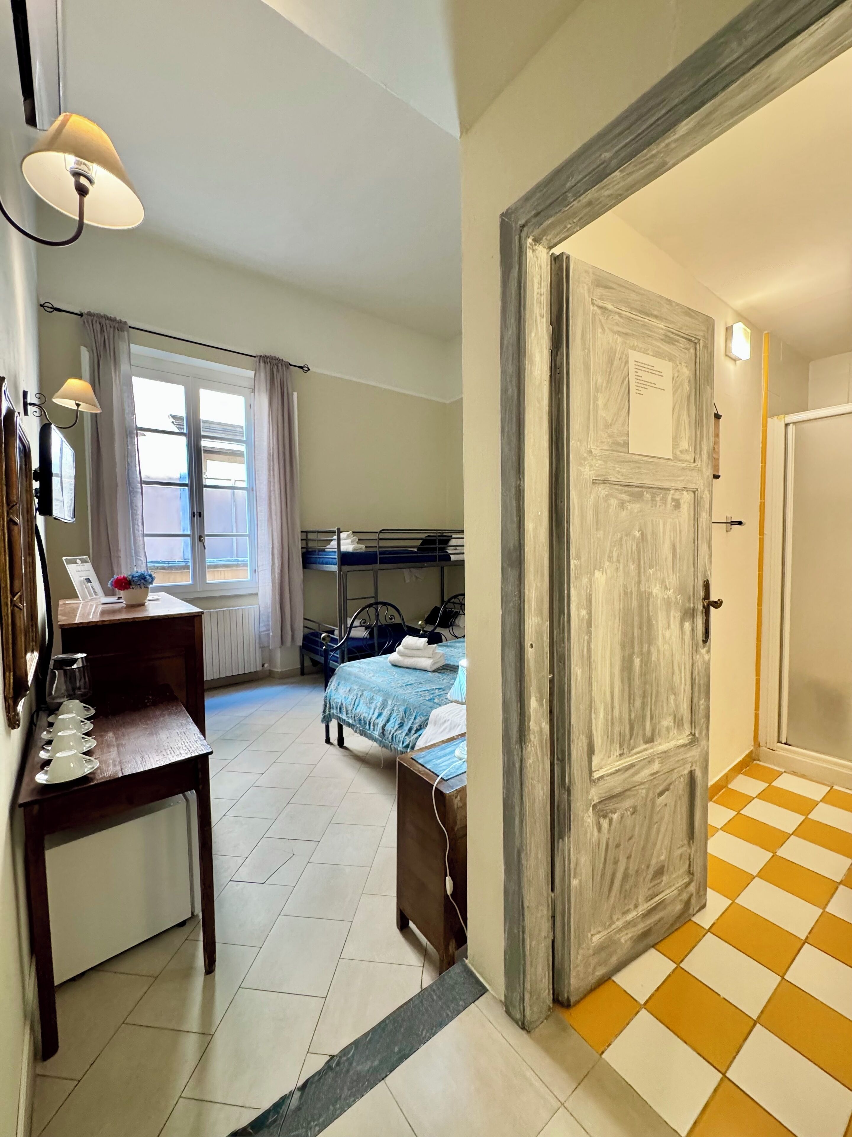 Una habitación con una cama, un escritorio con lámpara, una ventana con cortinas y una puerta con un letrero.