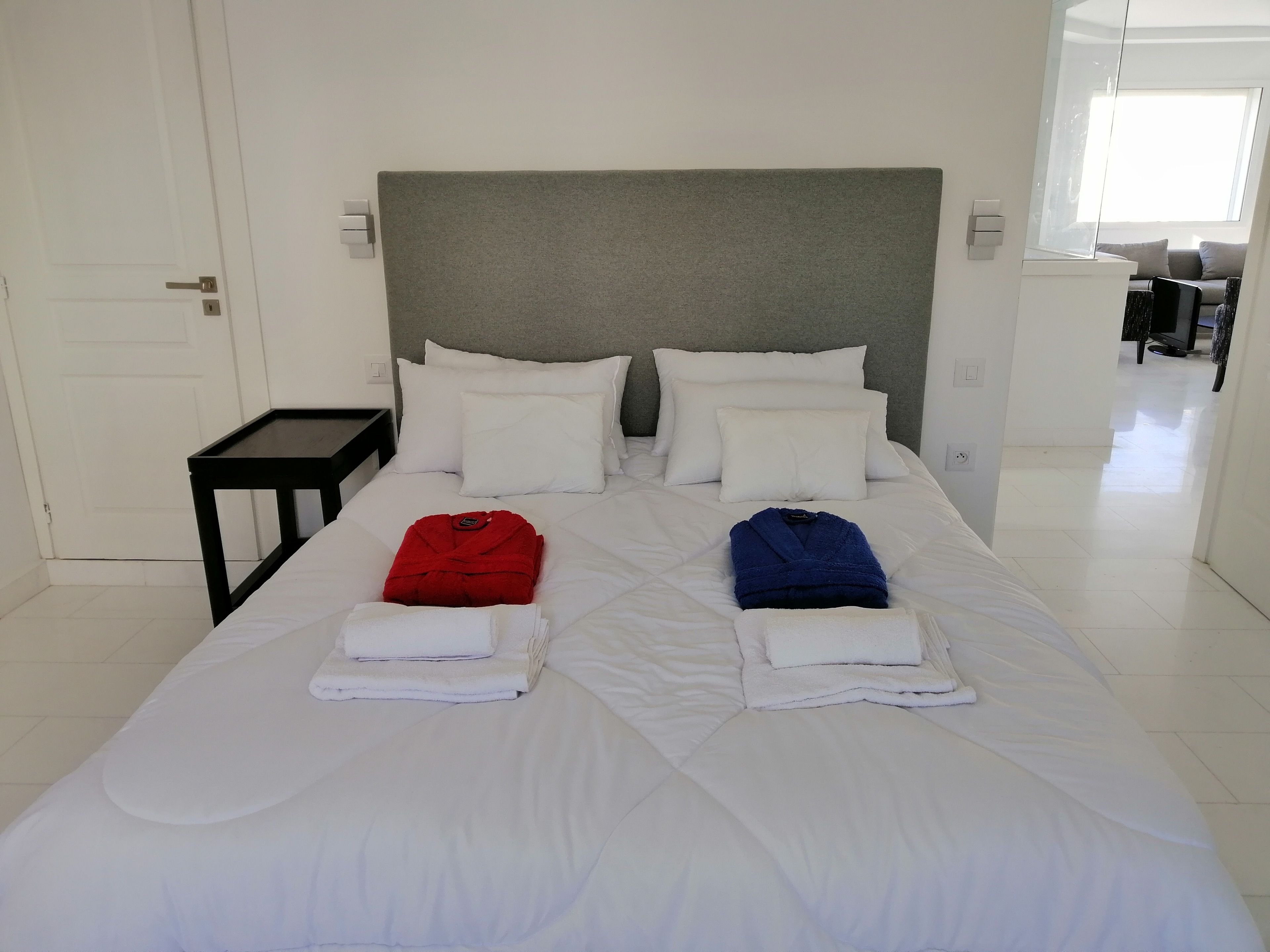 A neatly made bed with white bedding, two folded towels, and two red and blue bathrobes.