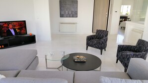A modern living room with a grey sofa, a black coffee table with decorative items, and two patterned armchairs. A television is on the left, and a radiator is on the wall. - Luxurious Apartment in le Palace (La Marsa)
