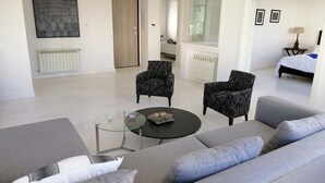 A modern living room with a grey sofa, two patterned armchairs, a glass coffee table, and a black side table. A bed with white bedding is visible in the background. - Luxurious Apartment in le Palace (La Marsa)