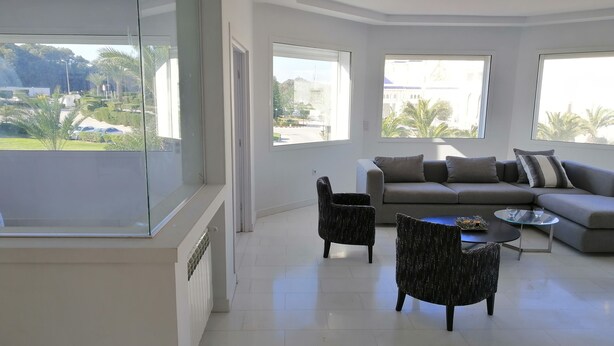 A modern living room with a grey sofa, two armchairs, and a glass coffee table. Large windows offer a view of a green landscape and buildings. - Luxurious Apartment in le Palace (La Marsa)
