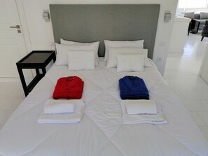 A neatly made bed with white linens, two folded towels, and two bathrobes in red and blue.