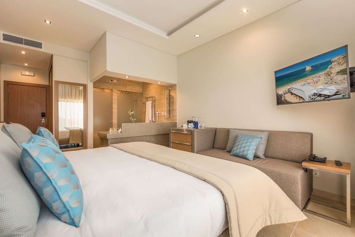 Een hotelkamer met een groot bed, een bank, een kleine tafel, een spiegel en een schilderij van een strandlandschap.