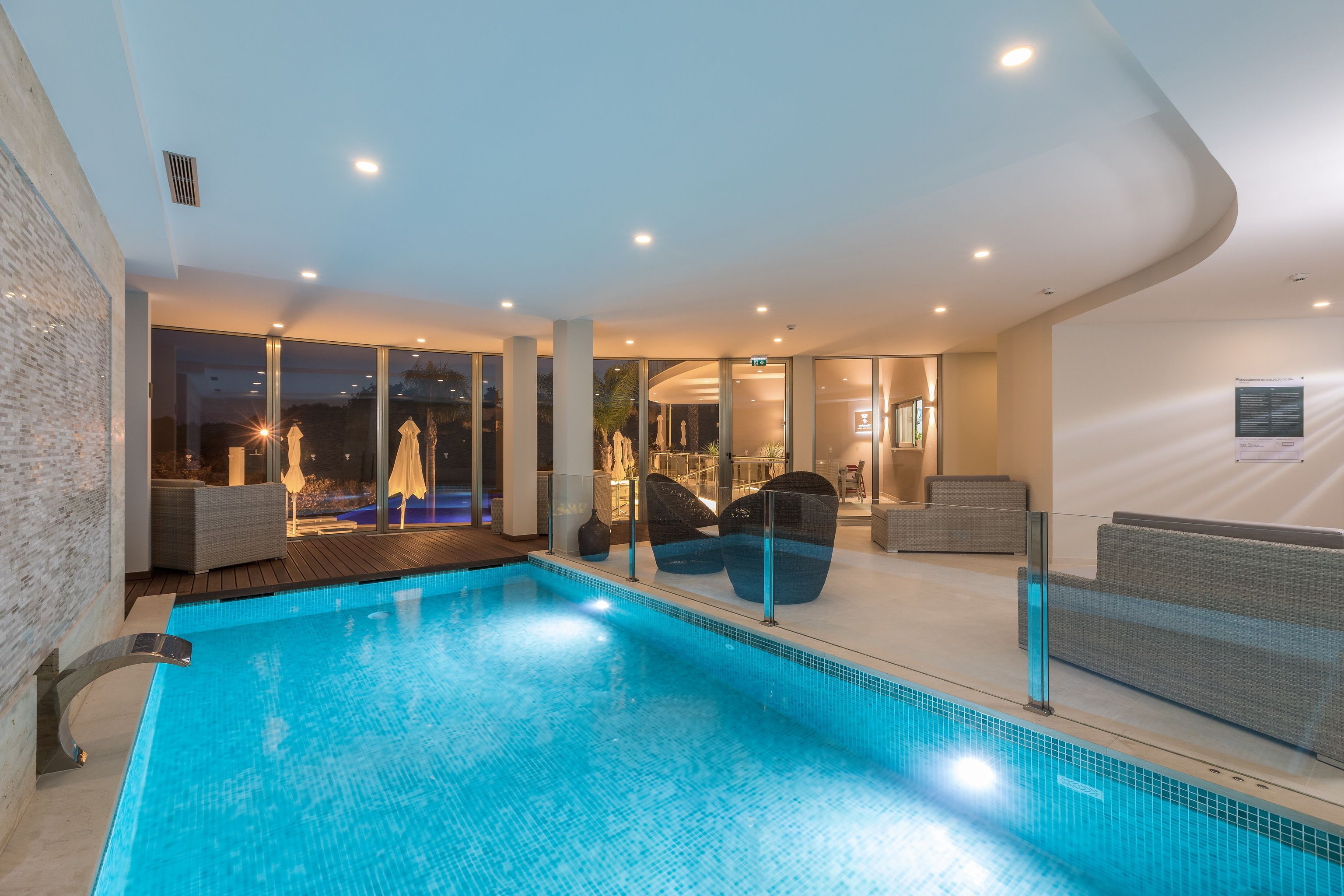 An indoor swimming pool with glass walls, modern seating, and recessed lighting.