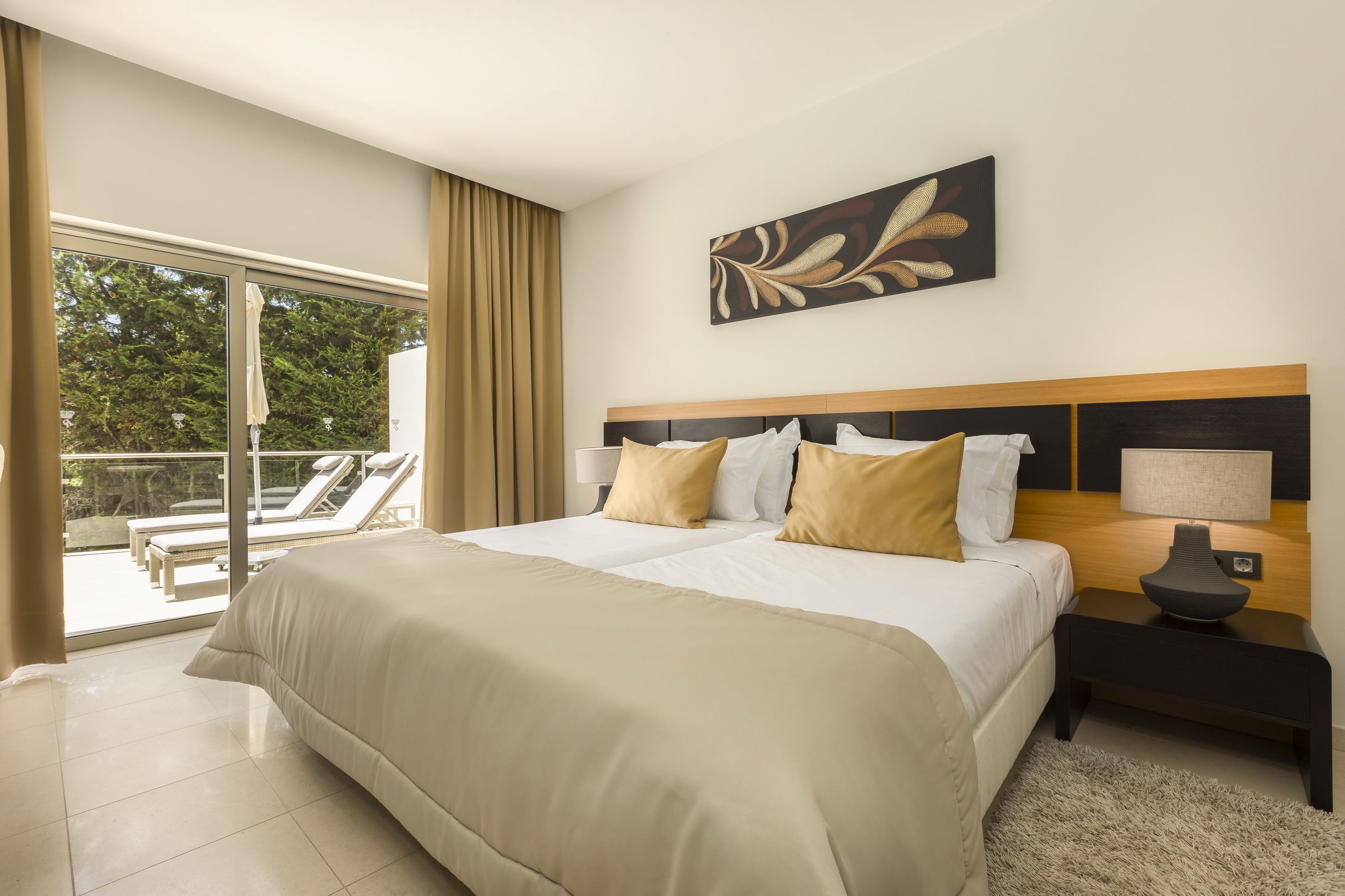 Habitación de hotel con una cama grande, una mesita de noche, una lámpara y vistas a un balcón con sillones de descanso.