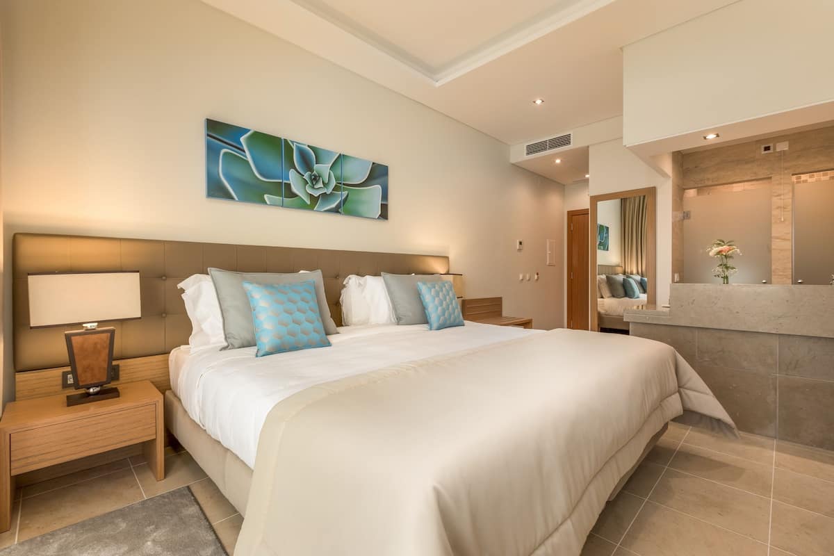 Een moderne hotelkamer met een groot bed, een nachtkastje met lamp, een kunstwerk aan de muur, en een badkamer met wastafel en spiegel.