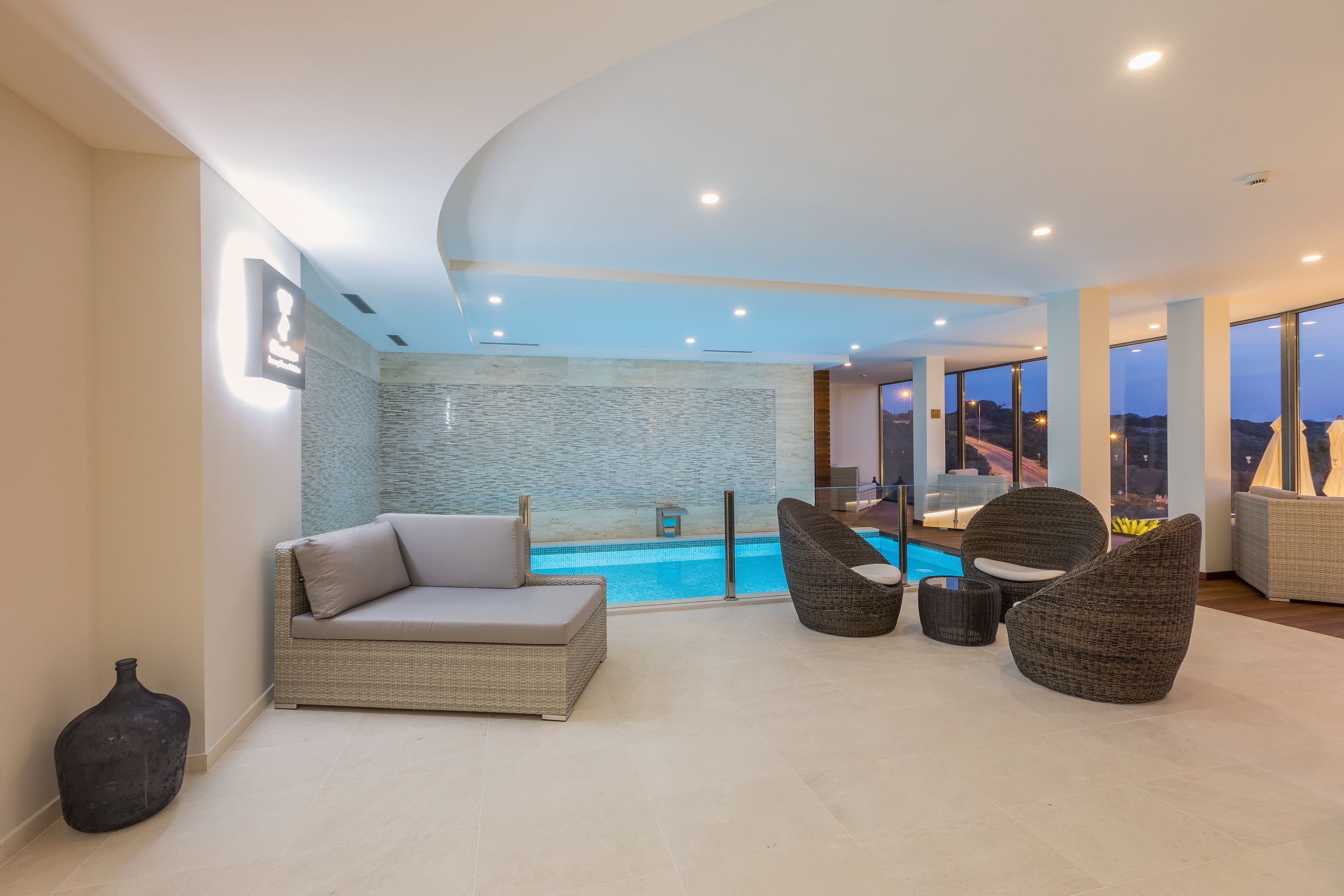 A modern indoor pool area with a sofa, two chairs, and a view of the pool through a glass wall.