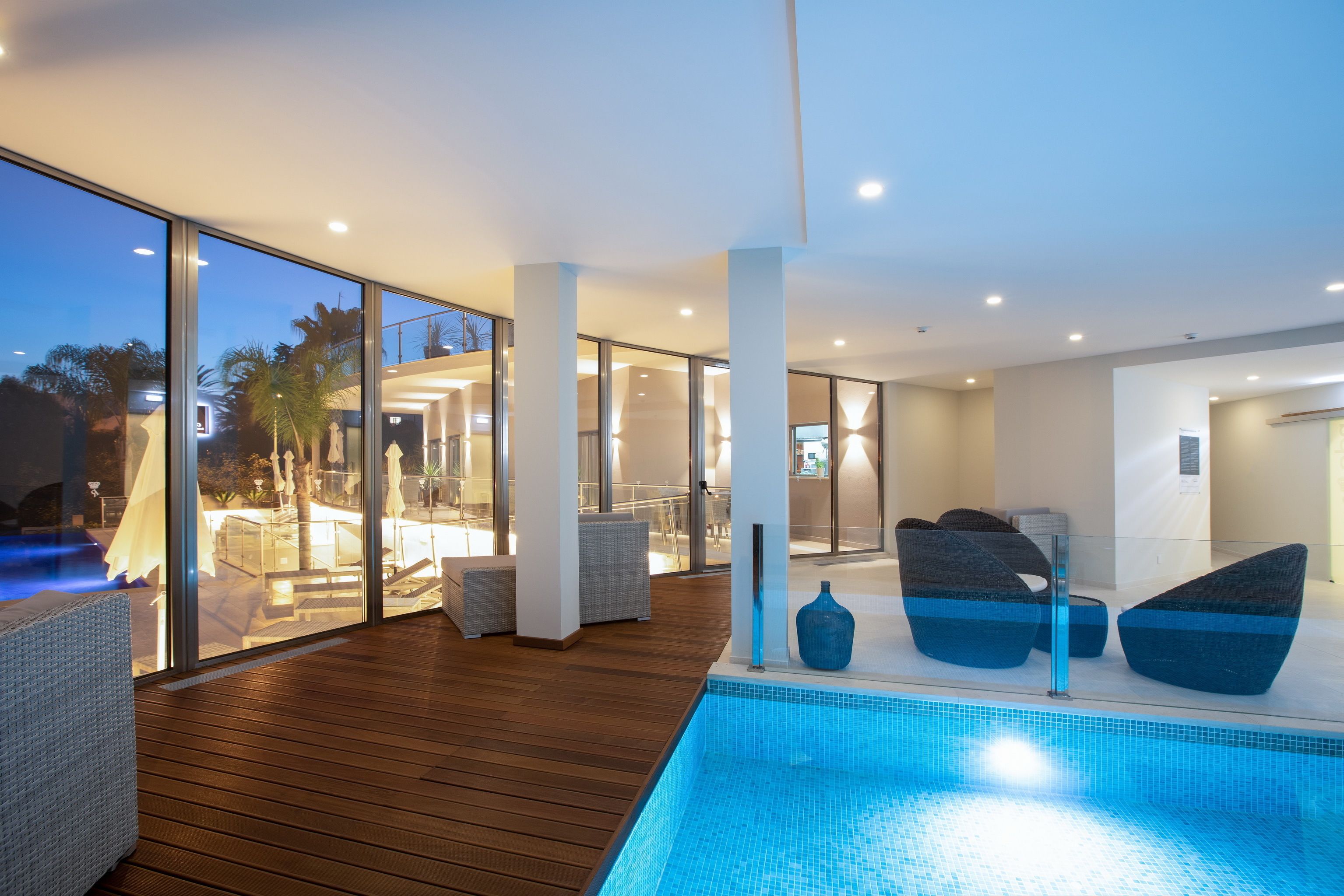 A modern indoor pool area with glass walls, wooden flooring, and contemporary seating.