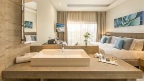 Ett modernt hotellrum med ett stort, rektangulärt handfat, en säng med flera kuddar, ett sängbord med toalettartiklar och ett stort fönster med gardiner.