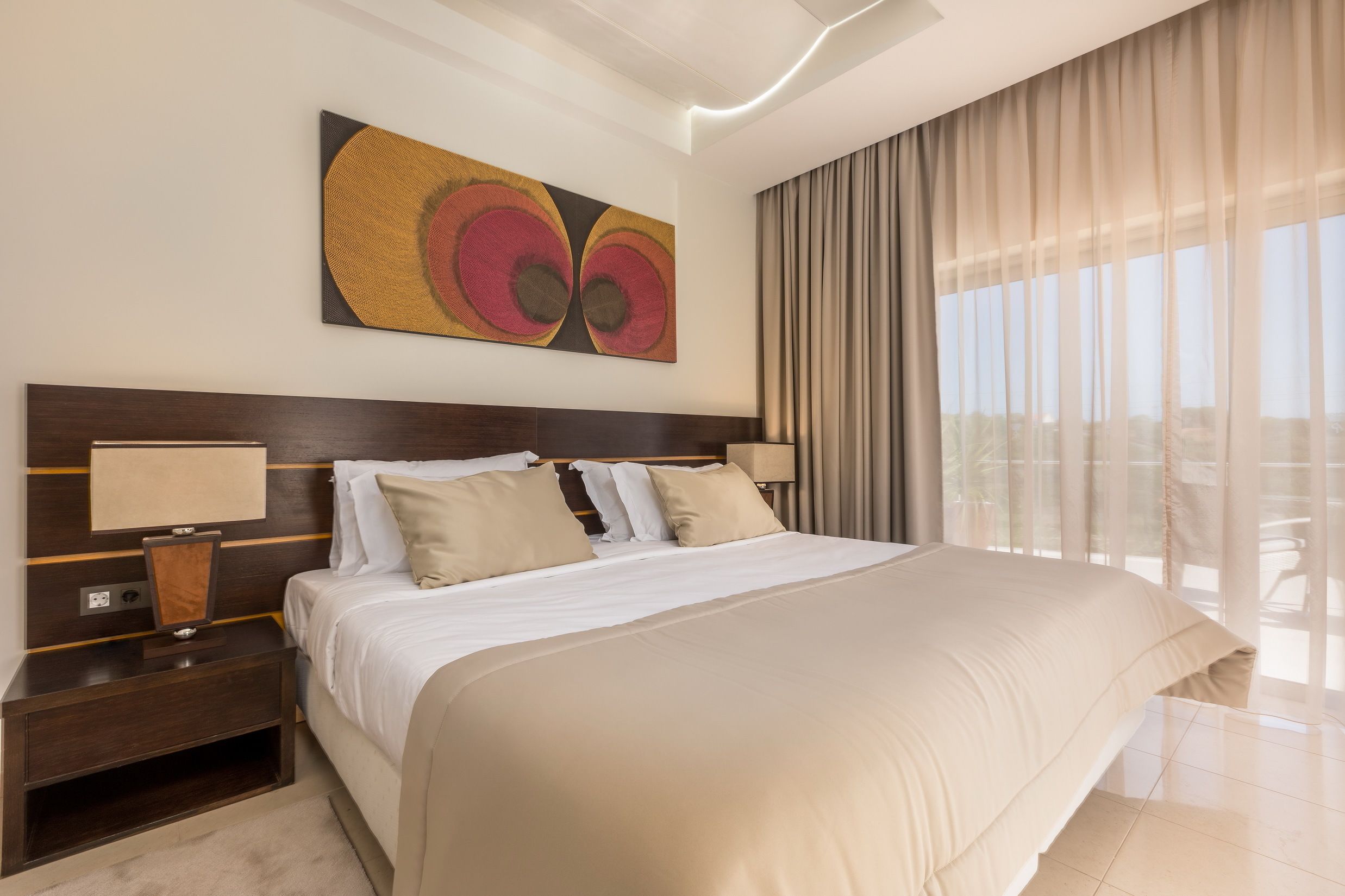A hotel room with a large bed, two bedside tables, a wall-mounted artwork, and a view through sheer curtains.