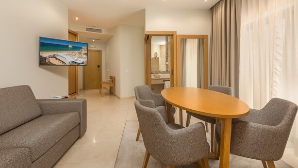 Una sala moderna con un sofá, una mesa de comedor con sillas, un televisor en la pared y vista al mar por la ventana.