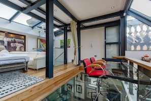 Quarto de hotel moderno com piso de vidro, cama, sofá e área de jantar com cadeiras vermelhas.