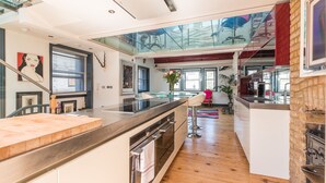 A modern kitchen with a central island, a dining area with bar stools, and a view of the living room through a glass ceiling. - The Museum Penthouse (London)