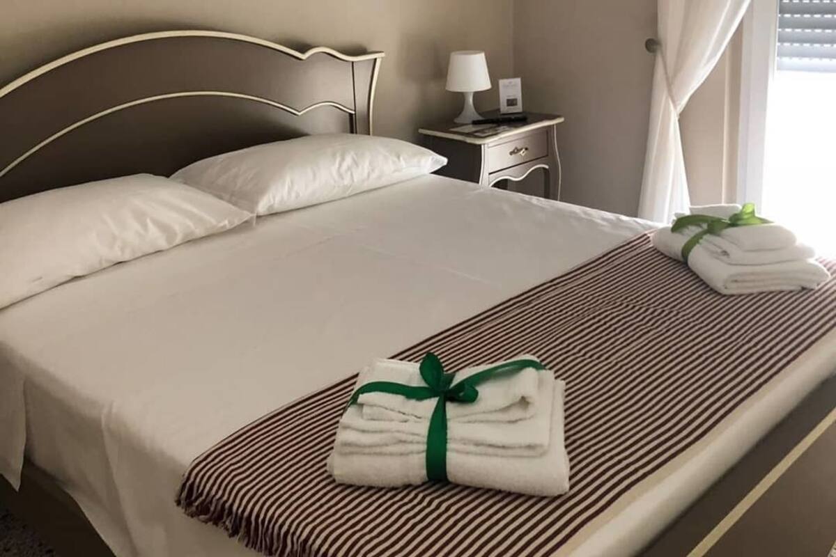 Un lit bien fait, avec des draps blancs et une couverture à rayures, une table de chevet avec une lampe et une fenêtre avec des rideaux.