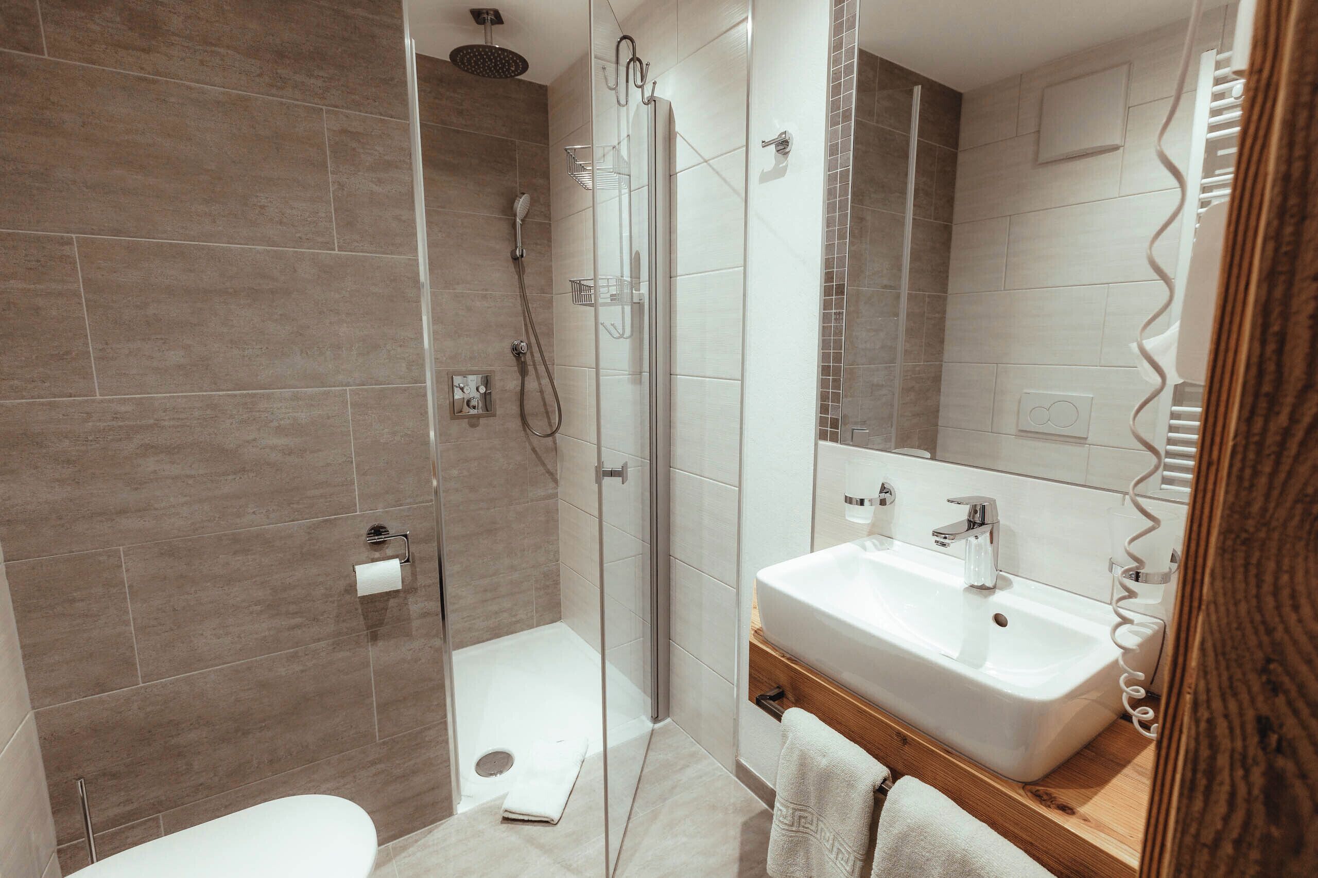 Un bany modern amb plat de dutxa envoltat de vidre, un lavabo blanc i un armari de tocador de fusta.