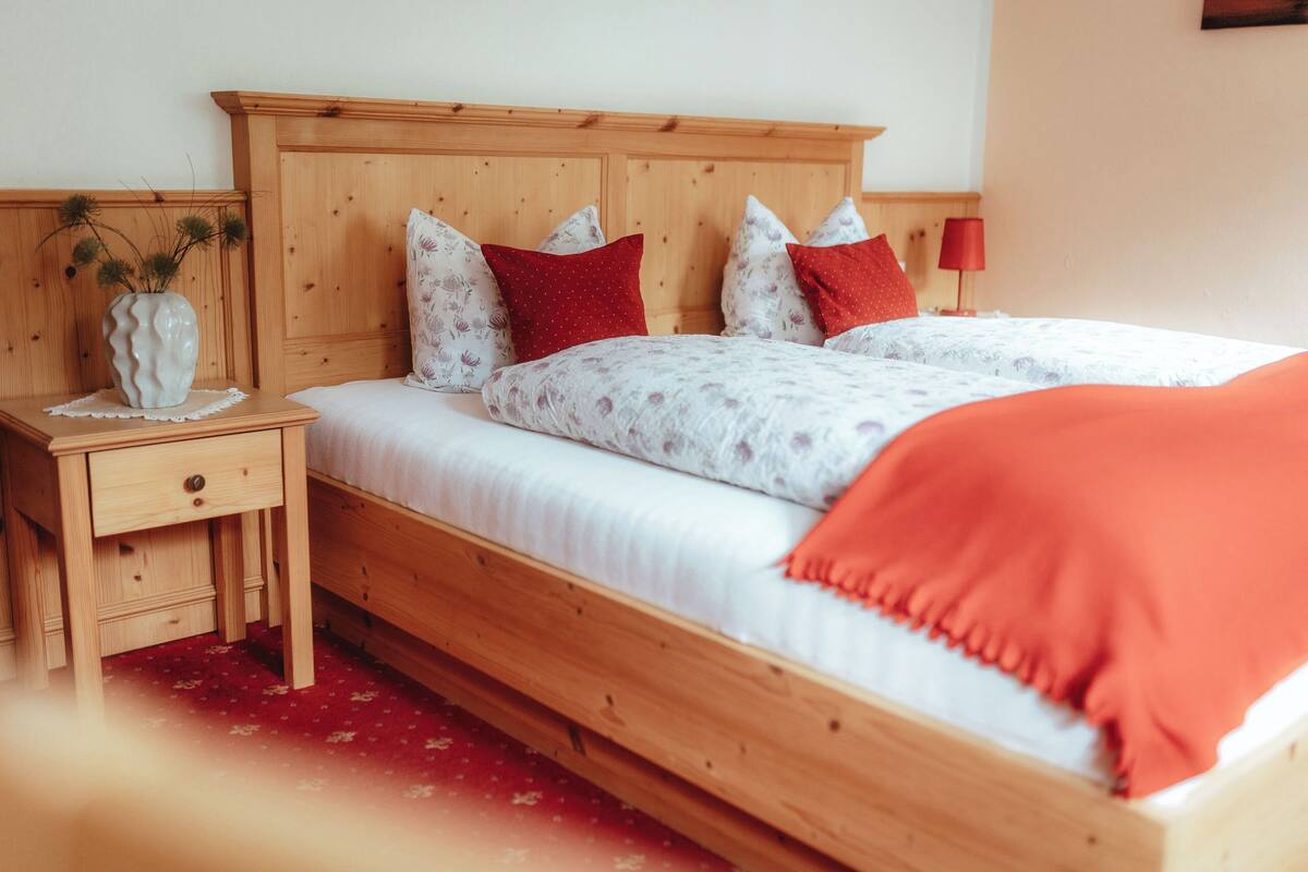 Un lit en bois avec une literie blanche et des oreillers rouges, une table de chevet avec un vase, et un tapis rouge.