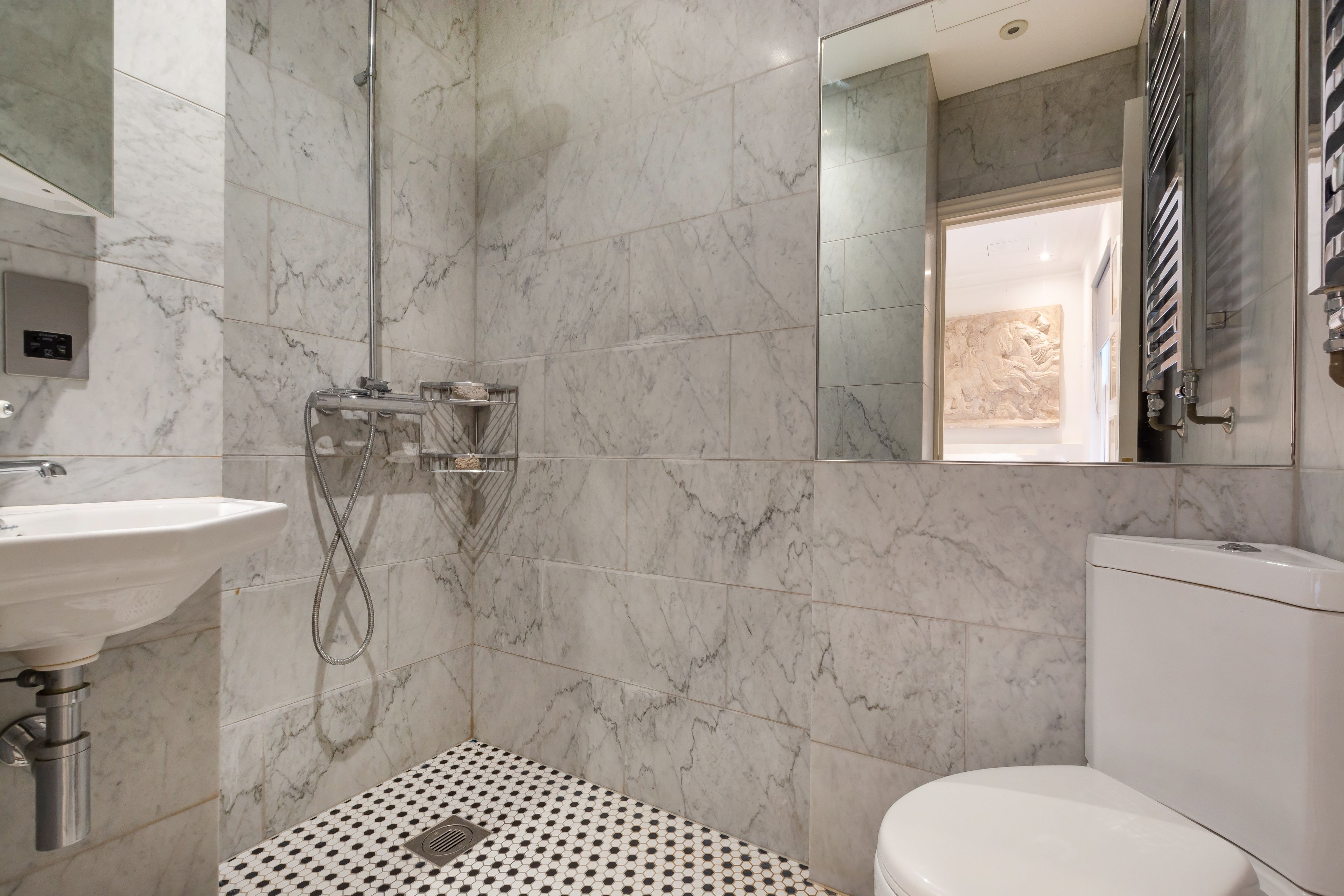 Un baño con piso de mármol, lavamanos blanco, inodoro y ducha con cabina de vidrio.