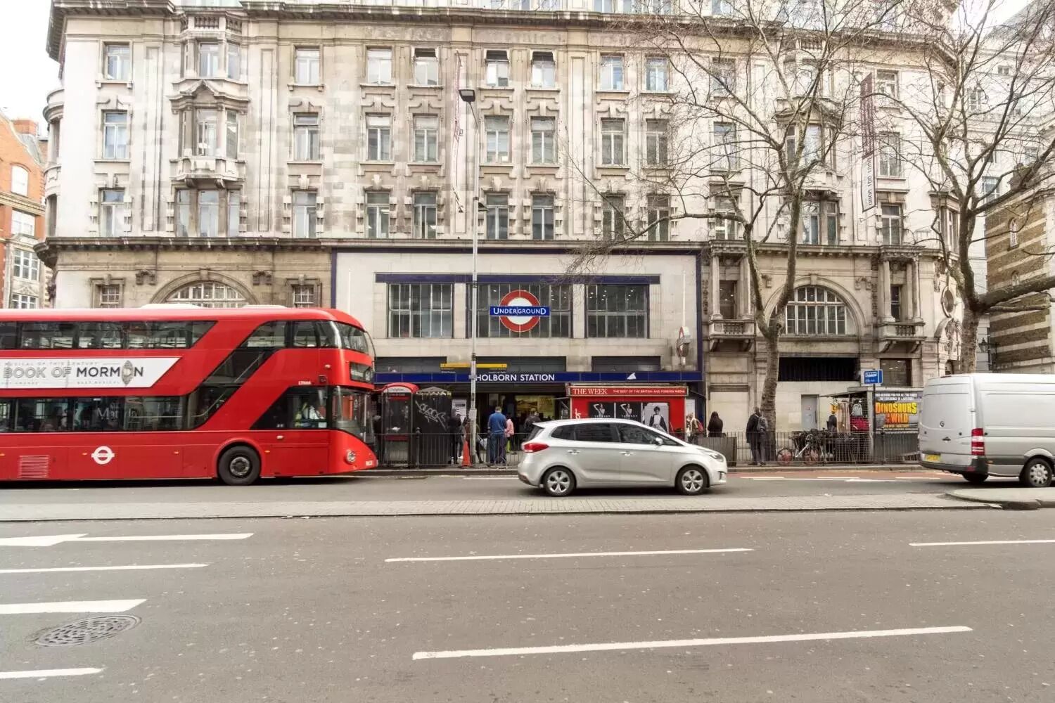 A red double-decker bus, a silver car, and a white van on a city street with a historic building in the background.