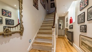 Escada com degraus de madeira e corrimãos brancos, ladeada por paredes decoradas com quadros e um vaso vermelho.