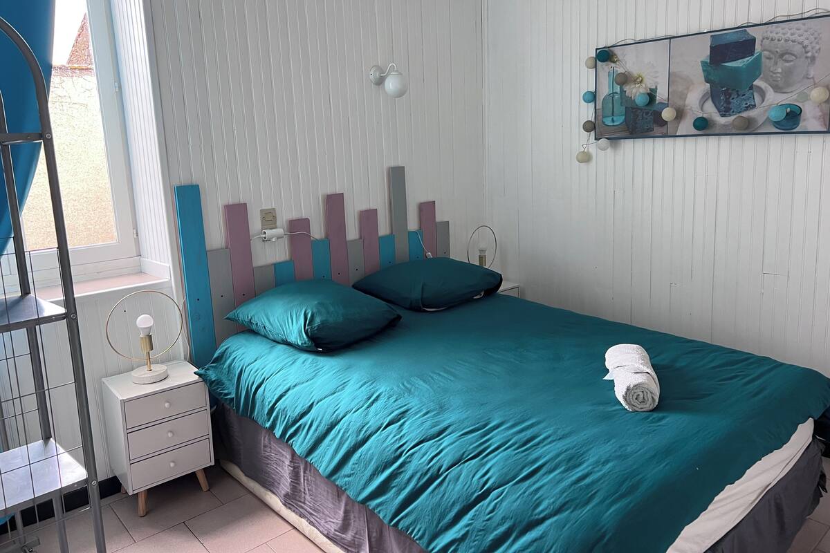 A bedroom with a teal bedspread, a white bedside table, a nightstand, a window with blinds, and a wall-mounted shelf with decorative items.