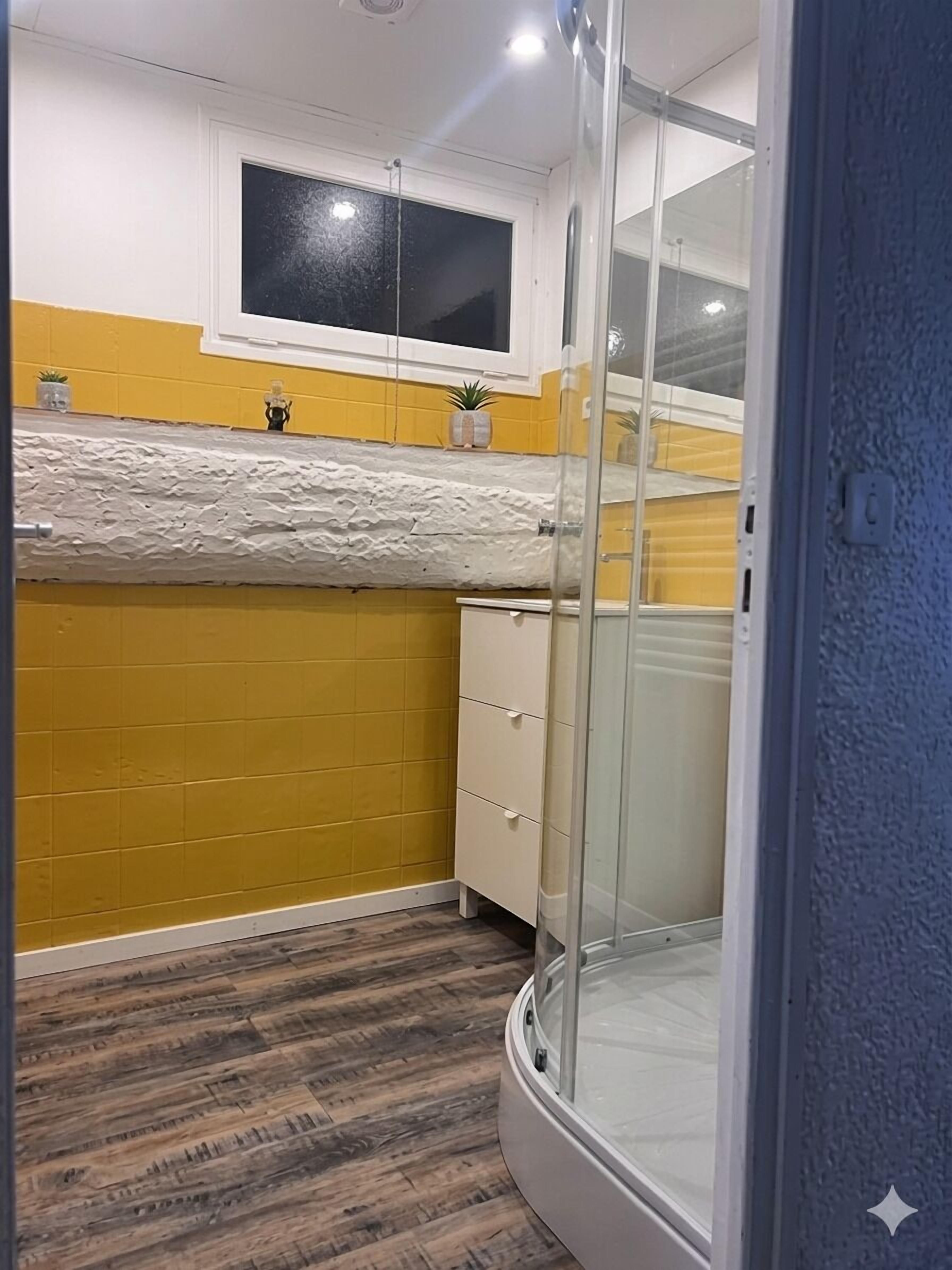 Un bany amb parets grogues, una banyera blanca i un plat de dutxa amb envàs de vidre.