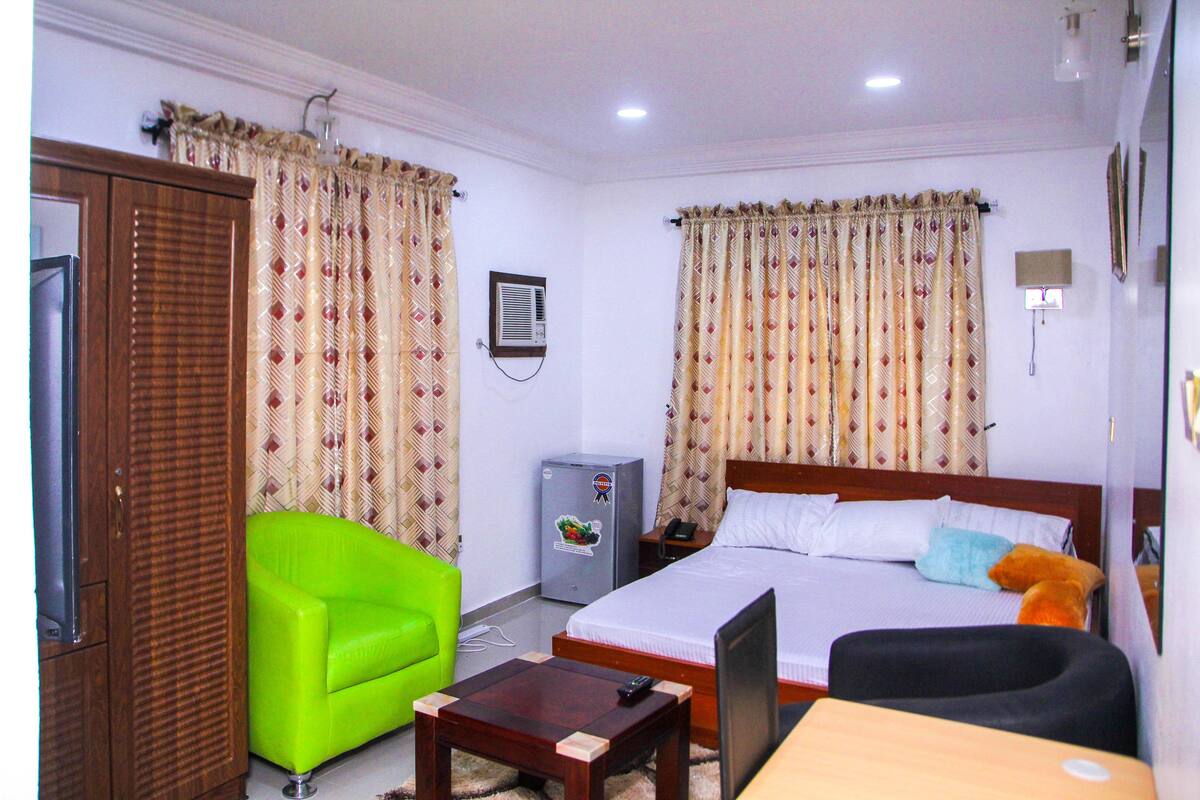 A hotel room with a bed, a green armchair, a wooden table, a desk, a television, a refrigerator, and curtains.