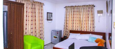 Una habitación de hotel con una cama, un sillón verde, una mesa de madera, un escritorio, un televisor, un refrigerador y cortinas.
