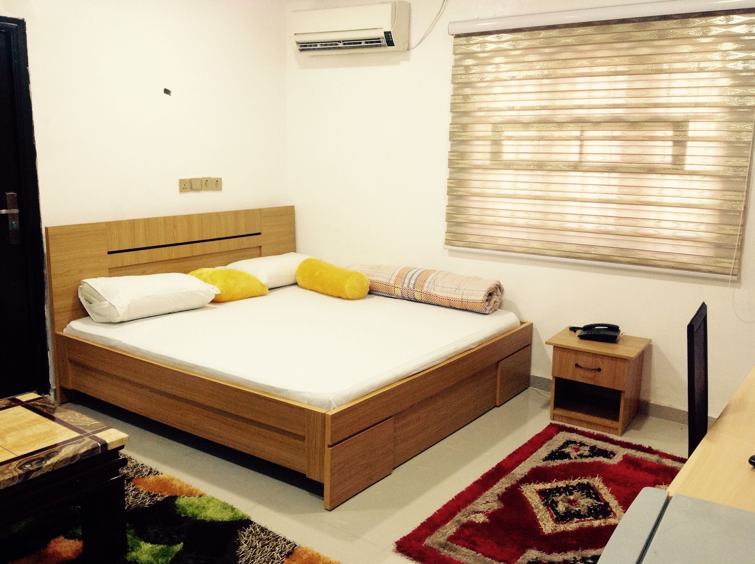 Un dormitorio con cama de madera, mesita de noche, un mueble pequeño, televisor y ventana con persianas.