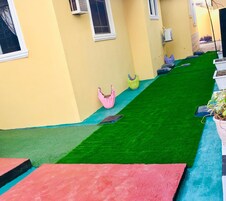 Un patio estrecho con césped artificial, un adorno rosa y amarillo, y una planta en maceta.