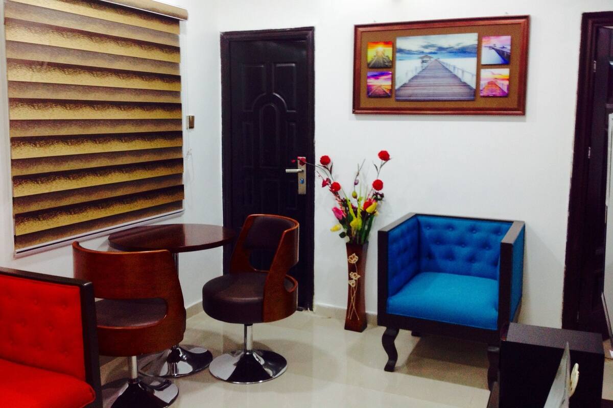 A room with a black door, a framed artwork, a vase with flowers, and two chairs.