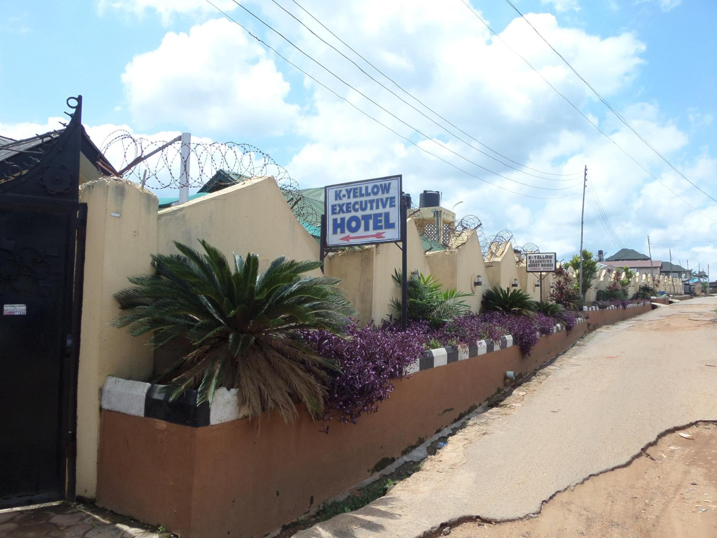 A K-Yellow Executive Hotel with a gated entrance, a signboard, and a landscaped area with plants.