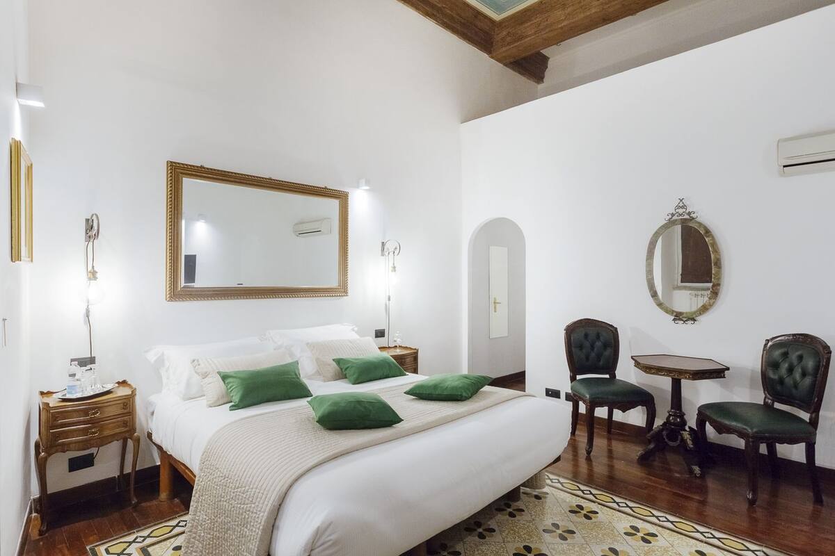 Habitación de hotel con una cama grande, dos mesitas de noche, un espejo, una pequeña mesa, dos sillas y una puerta.