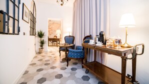 Ein Hotelzimmer mit einem sechseckig gemusterten Boden, einem Holztisch mit Lampe, einem Sessel und einem Schreibtisch mit Kaffeemaschine und Gläsern.